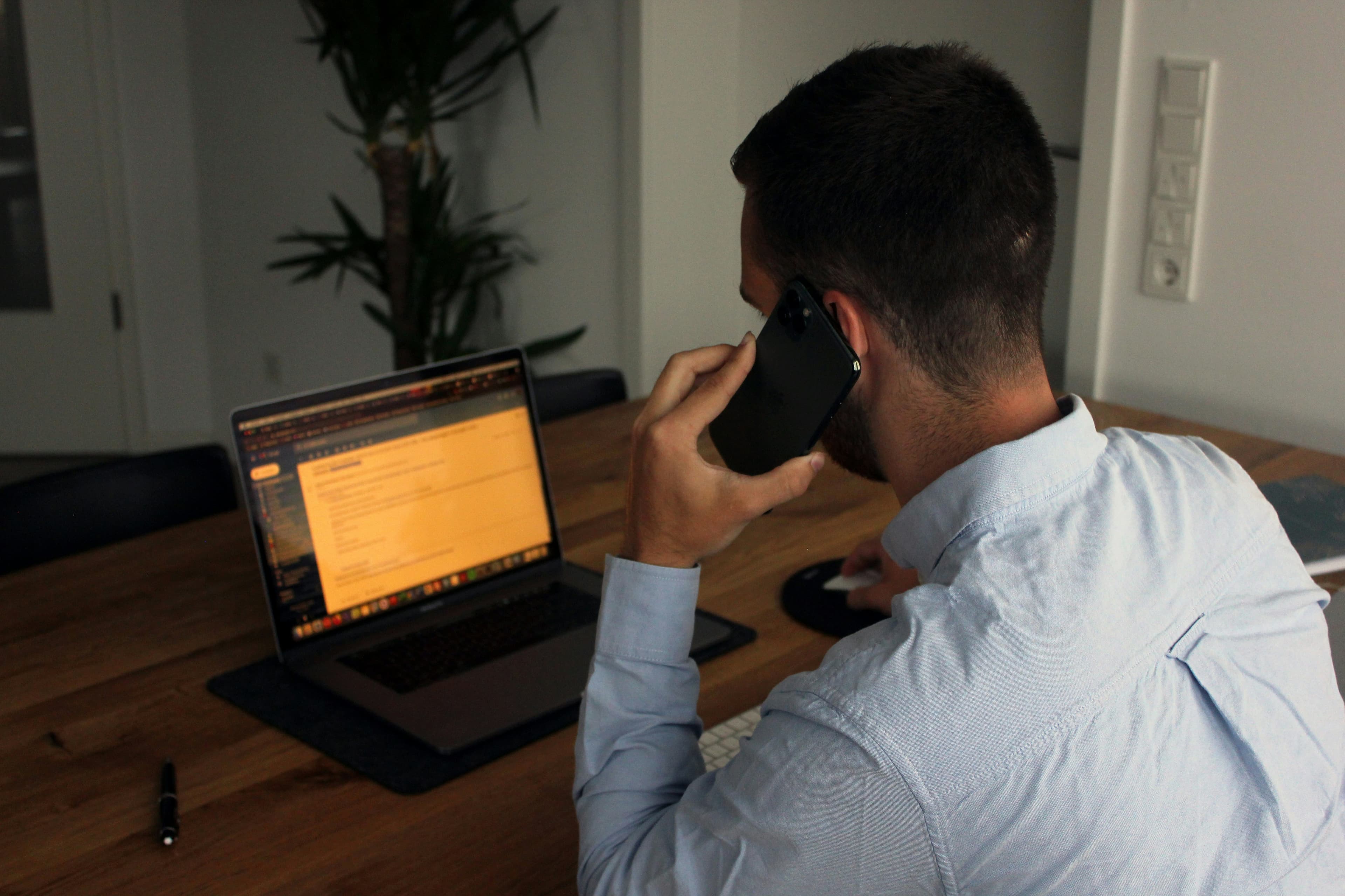 Man in blue shirt talking on a smartphone while working at a desk with laptop.