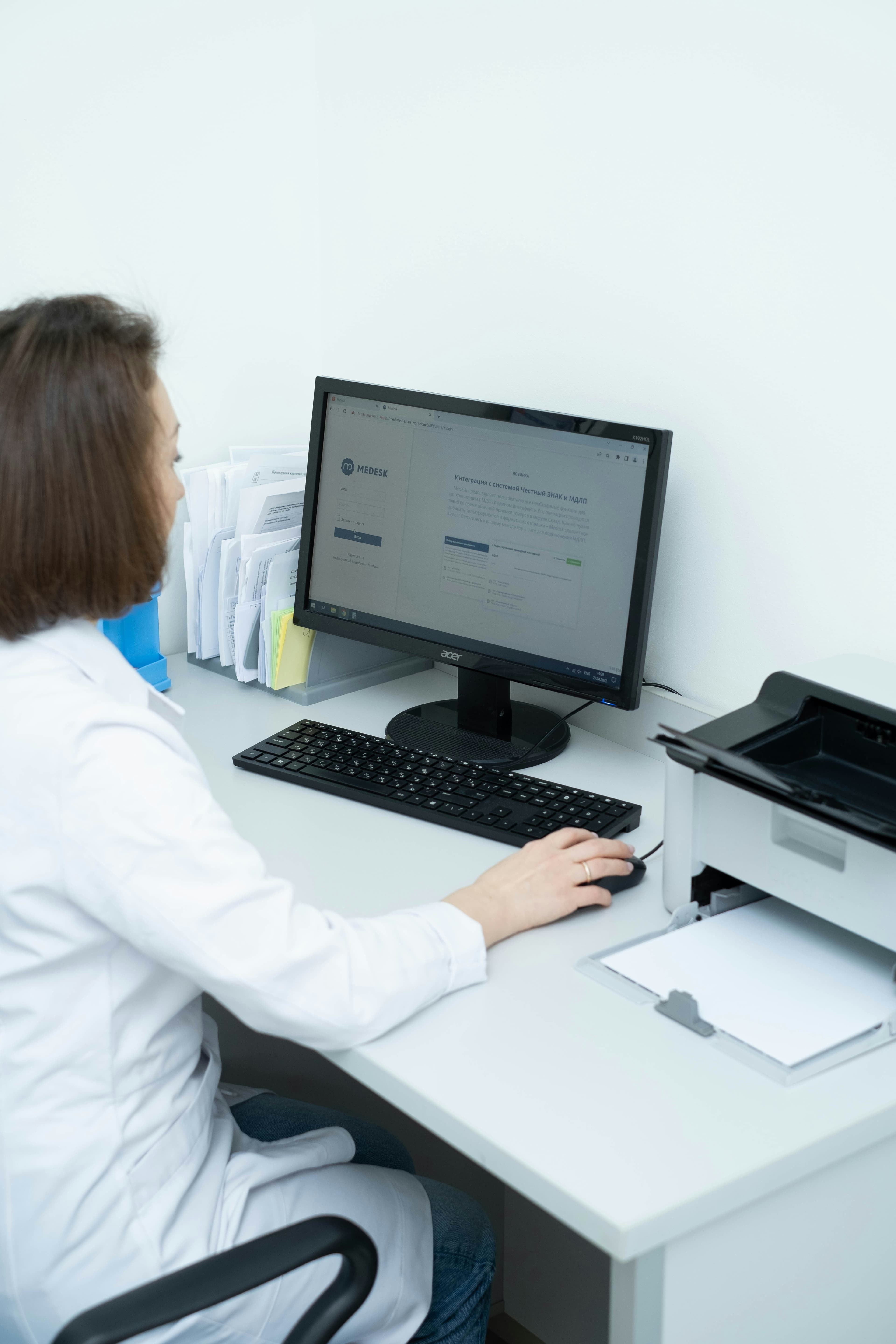 Healthcare professional in a white coat using a computer at a desk with a printer.