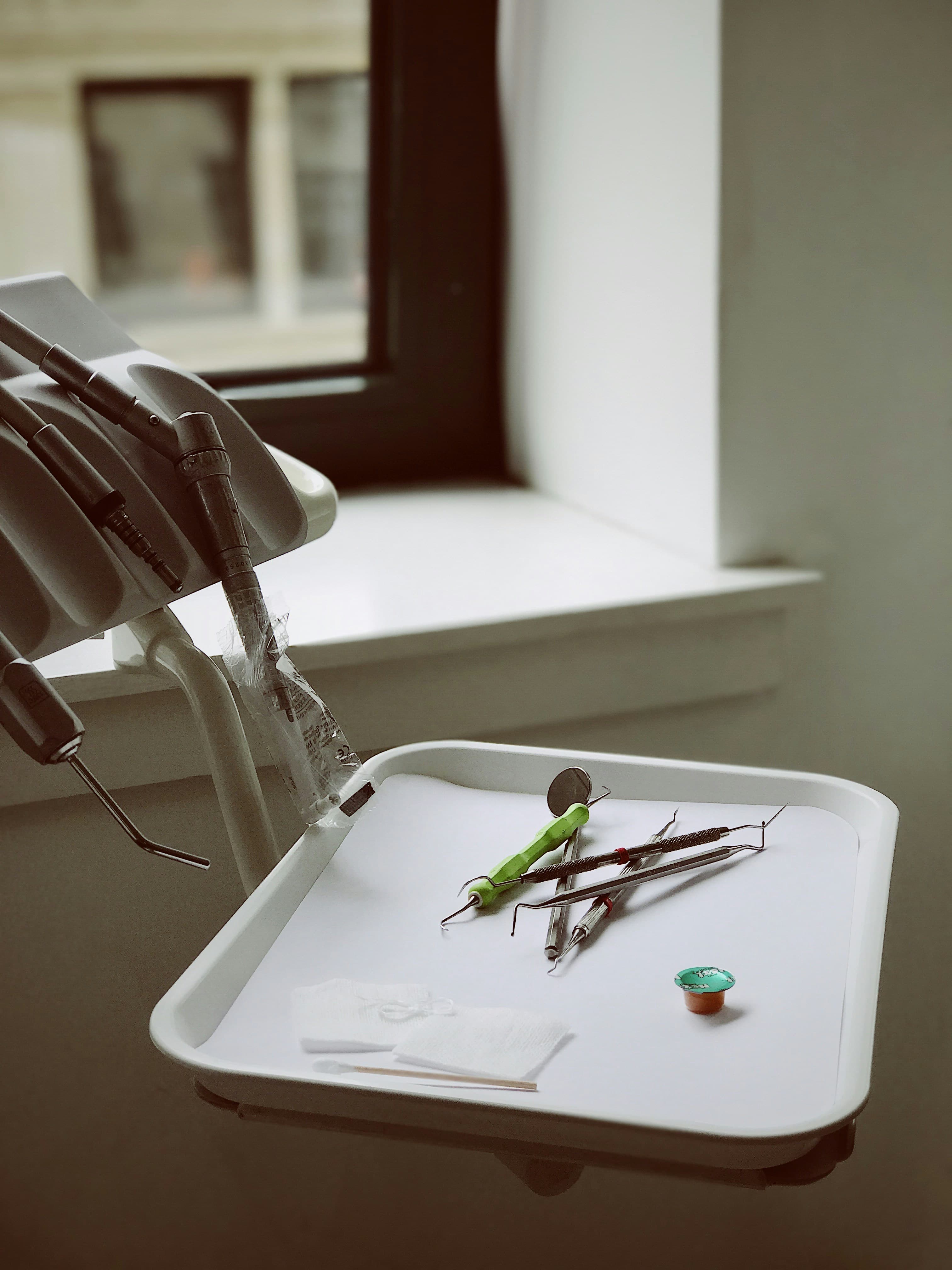 Dental instruments on a white tray beside a dental handpiece unit near a window.