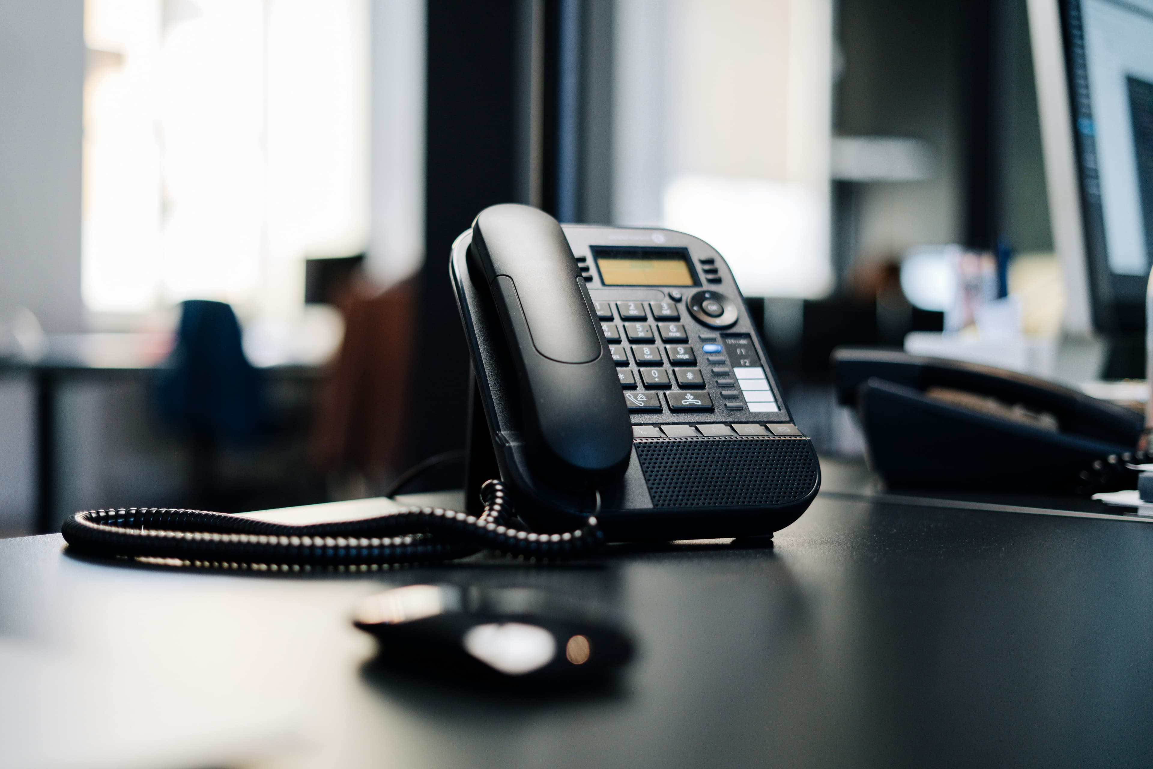 Black office desk phone with coiled cord on a dark desk in a blurred office.