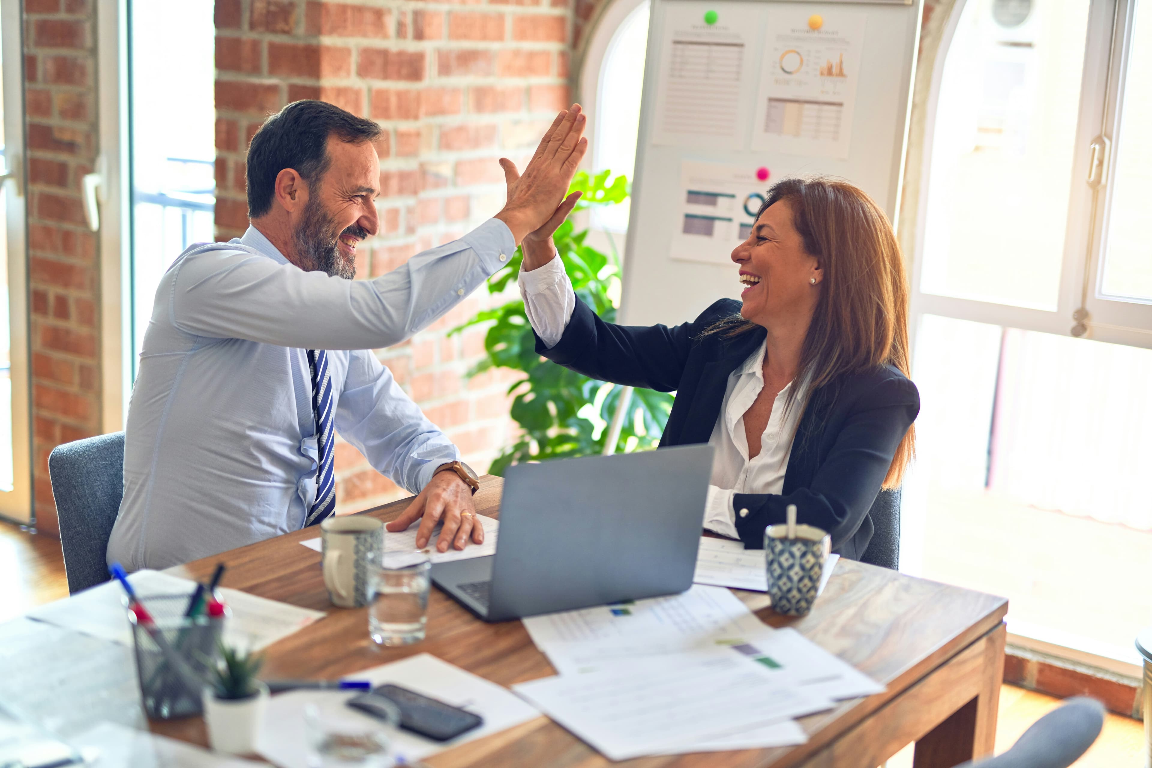 Two smiling professionals high-five over a laptop at a desk in a bright office.
