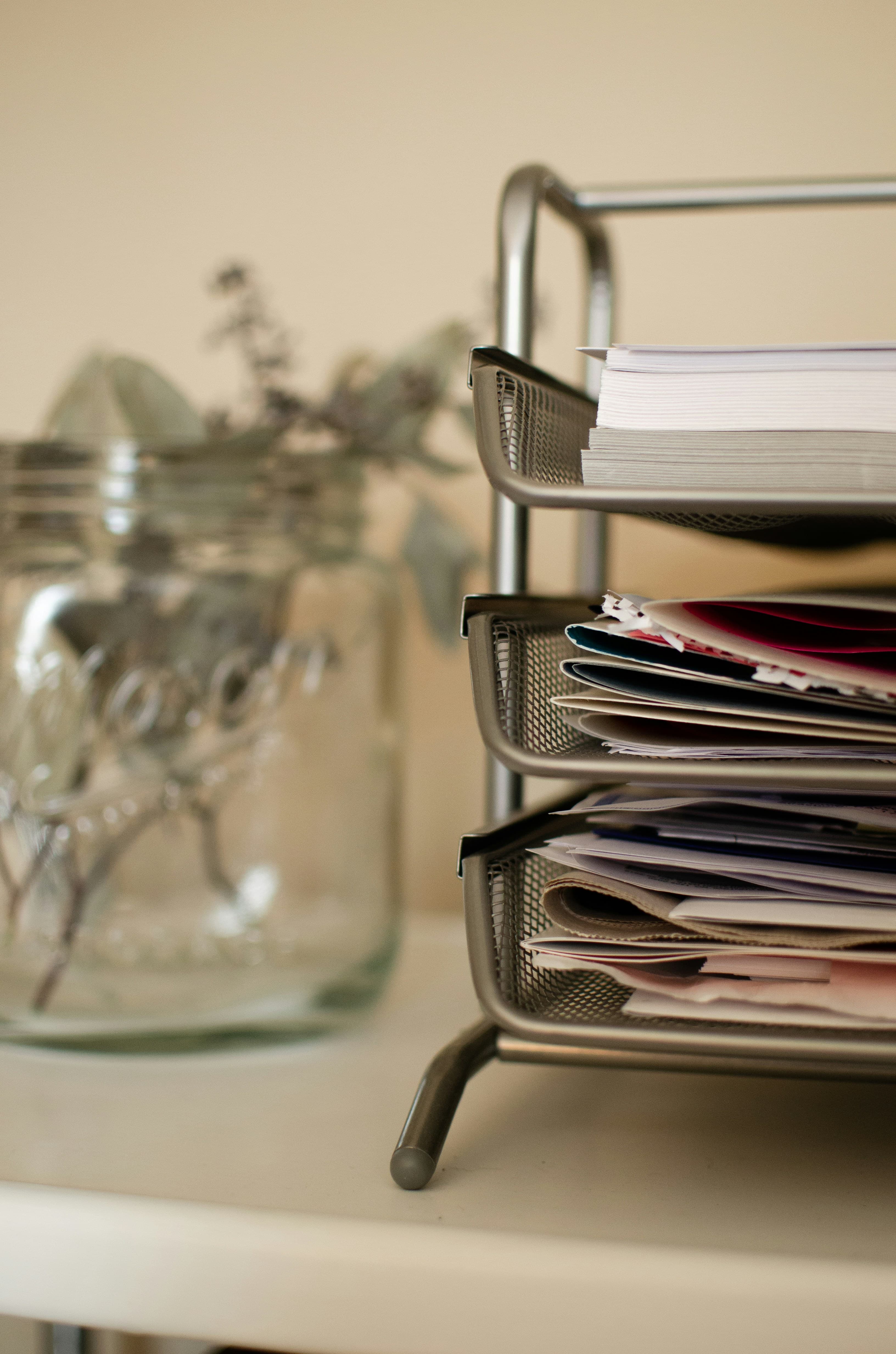 Metal mesh desk organizer overflowing with papers and mail beside a blurred glass jar.