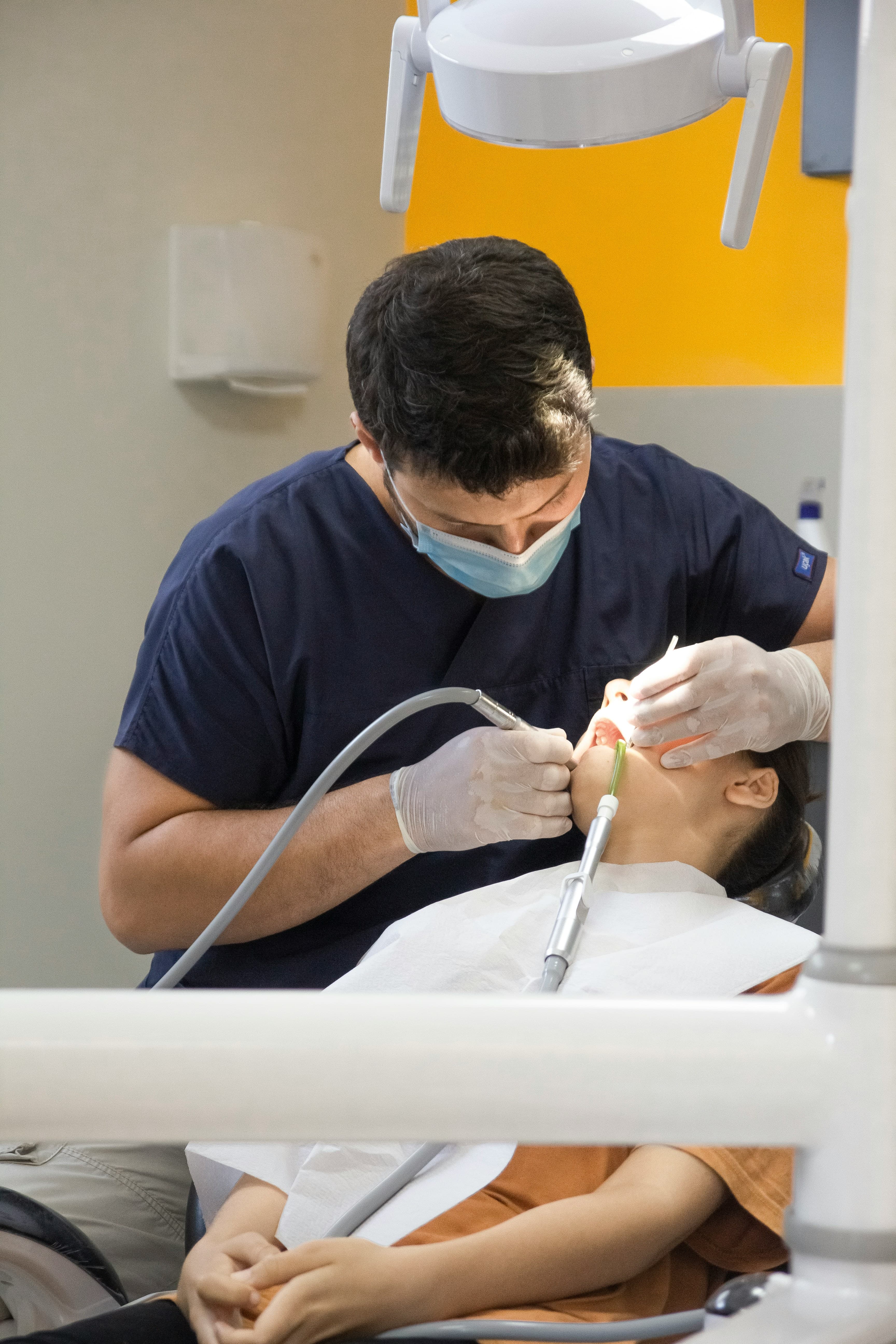 Male dentist in blue scrubs and mask performs a procedure on a young patient.